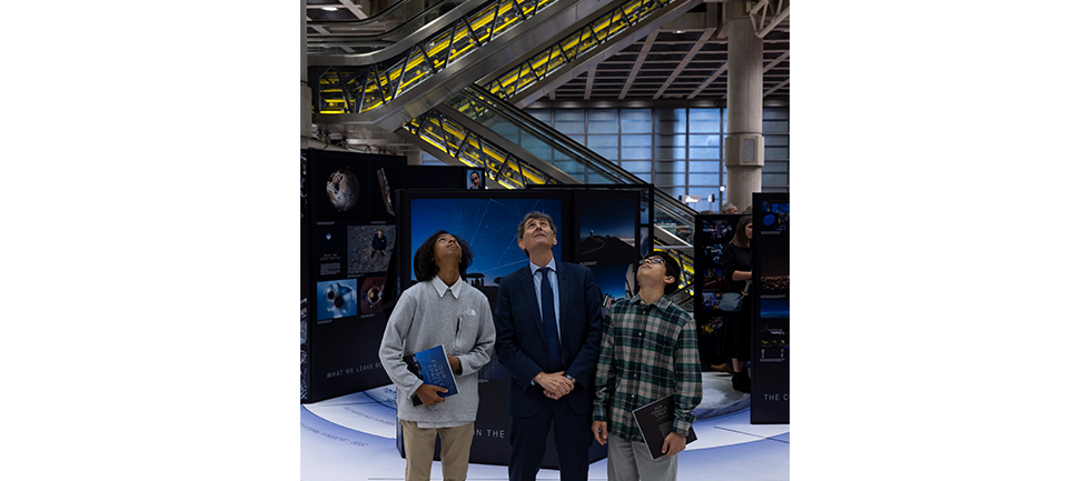 Image of competition winners and max alexander standing next to each other look up to sky, they are standing in front of the exhibition with a background of the escalators in the Lloyd's underwriting room